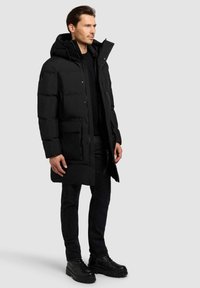 Zwarte puffy parka met capuchon, voorzien van een knoopsluiting aan de voorkant, twee grote zakken aan de voorkant en een gestructureerde stof. Gecombineerd met donkere jeans en laarzen.