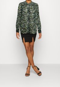 Blusa nera trasparente con un motivo floreale, maniche lunghe e chiusura con bottoni. Abbinata a una gonna nera aderente con fessure laterali e tacchi.