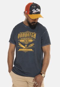 Camiseta de algodón azul marino con diseño gráfico amarillo de temática de barbería, llevada con una gorra multicolor con acentos de palmera.