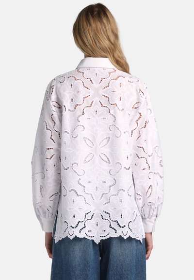 Frau trägt eine weiße langärmlige Bluse mit filigraner floraler Cut-out-Stickerei, kombiniert mit blauen Denim-Jeans, von hinten gesehen.