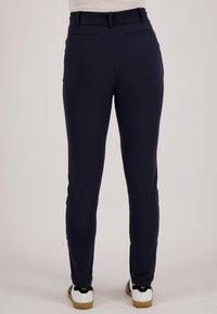 Persoon die nauwsluitende donkerblauwe broek en witte sneakers draagt, staand tegen een effen lichte achtergrond, gezien van achteren.