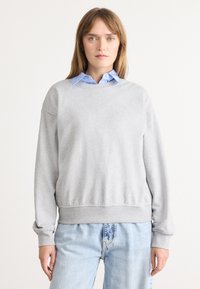 Felpa grigia chiaro con polsini e orlo a costine, scollatura rotonda, texture morbida, indossata sopra una camicia a collo blu. Sottile logo CK sul petto.