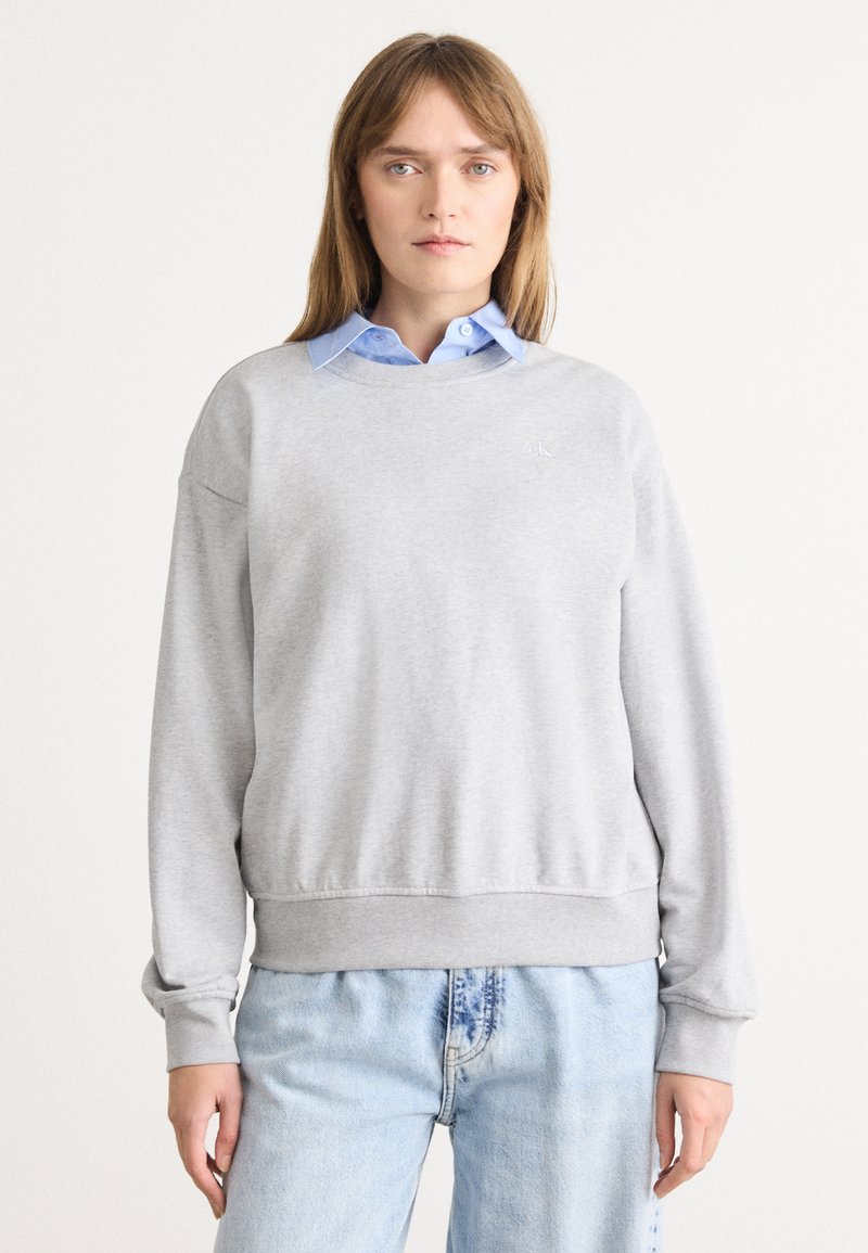 Felpa grigia chiaro con polsini e orlo a costine, scollatura rotonda, texture morbida, indossata sopra una camicia a collo blu. Sottile logo CK sul petto.