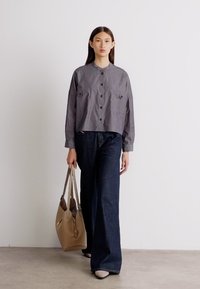 Grijs gestreepte cropped shirt met knopen aan de voorkant, gecombineerd met donkere wijdlopende jeans. Model draagt een beige handtas en staat tegen een neutrale achtergrond.