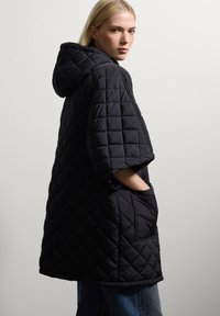 Schwarzer gesteppter Poncho mit Kapuze, breiten Ärmeln und einer Fronttasche. Der Stoff hat eine strukturierte Oberfläche und eine lockere Passform.