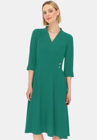 Turkusowa sukienka w kształcie kopertowym z rękawami o długości trzech ćwierci, dekoltem w kształcie litery V oraz rozkloszowaną spódnicą. Posiada dwa dekoracyjne guziki w talii, wykonane z gładkiego materiału.
