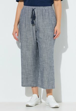 Kobieta ubrana w luźne szare szerokie spodnie do połowy łydki w paski z granatowym sznurkiem i białe trampki, stojąca przy białej ścianie.