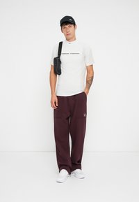 White Nike Tech t-shirt with black logo, maroon loose-fitting pants with pockets, black crossbody bag, black cap, and white sneakers.