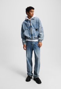 Denimjas met elastische taille, voorzakken en ritssluiting, gecombineerd met lichtblauwe jeans en zwarte chunky schoenen. Witte hoodie eronder.