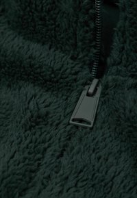 Ciemnozielony materiał z polaru o miękkiej fakturze, z szarym suwakiem zakończonym okrągłym uchwytem, który ułatwia chwytanie i użycie.