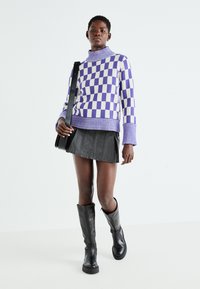 Pulover cu guler înalt, cu un model în carouri violet și crem, cu manșete și tiv ondulat, asortat cu o fustă plisată neagră și cizme negre până la genunchi.