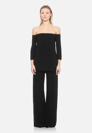 Frau trägt ein schwarzes Langarm-Oberteil mit schulterfreiem Ausschnitt und weit geschnittene schwarze Hose, steht vor einem schlichten hellen Hintergrund.