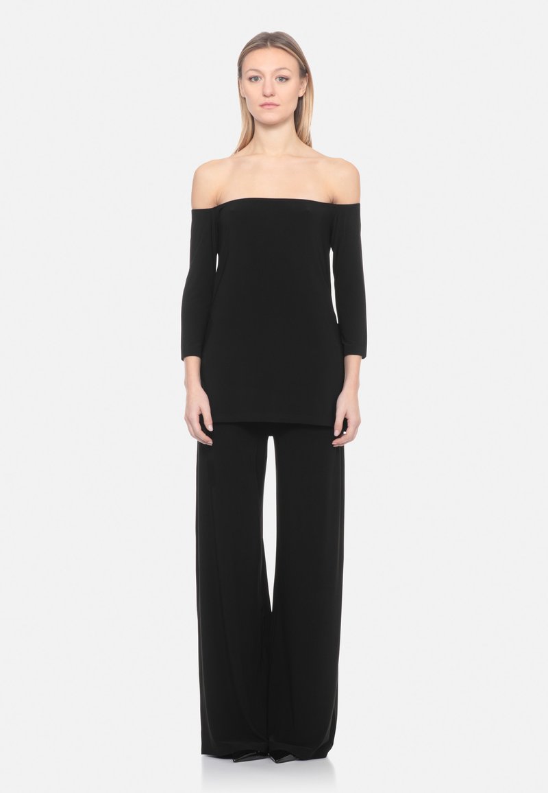 Vrouw draagt een zwart off-shoulder top met lange mouwen en zwarte broek met wijde pijpen, staand tegen een effen lichte achtergrond.