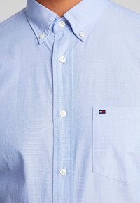 Hellblaues Button-Down-Hemd aus Baumwolle. Es verfügt über einen strukturierten Kragen, weiße Knöpfe und ein kleines Logo auf der Brusttasche.