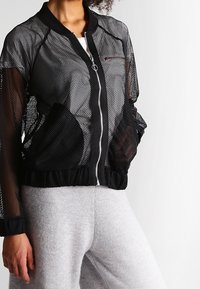 Modell som bär en svart mesh-bomberjacka över en vit topp, tillsammans med ljusgråa sweatpants, framför en neutral vit bakgrund.