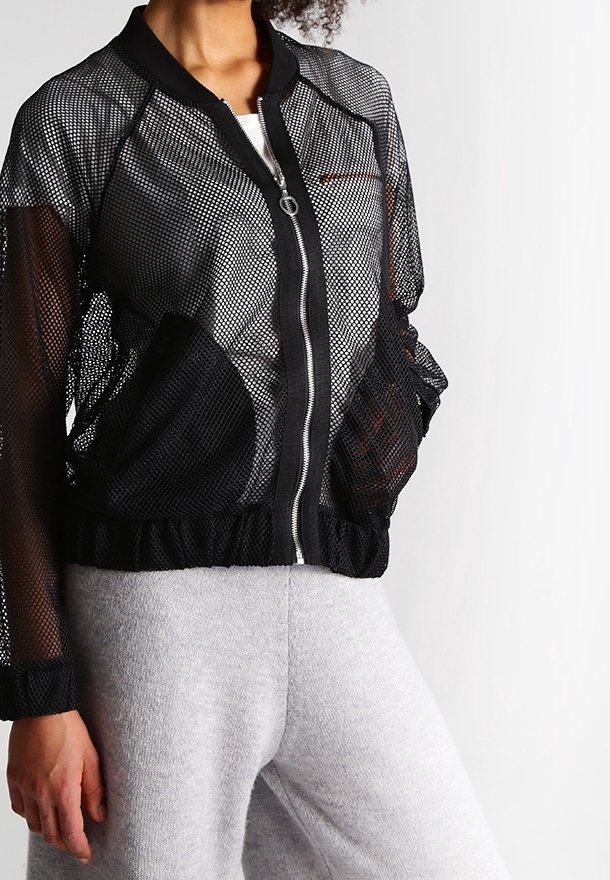 Modell som bär en svart mesh-bomberjacka över en vit topp, tillsammans med ljusgråa sweatpants, framför en neutral vit bakgrund.