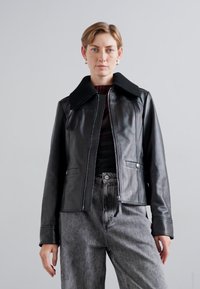 Black leather jacket with ribbed collar, front pockets, and contrasting stitching. Paired with high-waisted gray denim pants. Smooth texture.