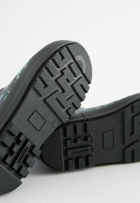 Suole di scarpe in gomma nere con scanalature profonde e un contrassegno di misura 12, posizionate su uno sfondo chiaro e semplice.