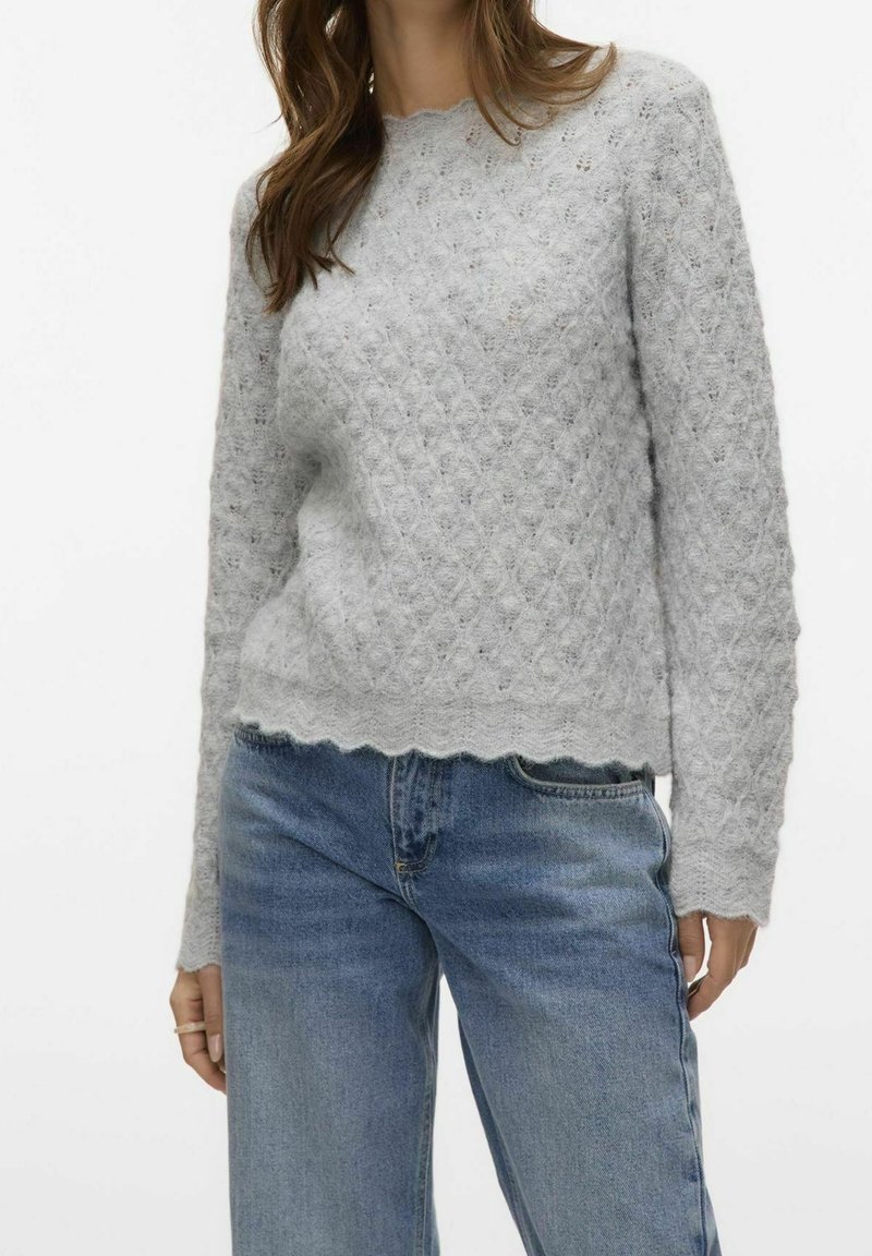 Femme portant un pull tricoté gris clair texturé avec des bords festonnés, associé à un jean bleu taille haute sur un fond uni.