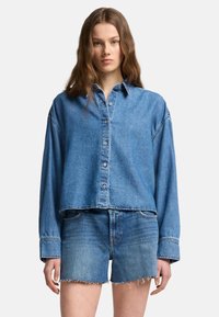 Camicia in denim azzurro chiaro con bottoni, dal design cropped e maniche lunghe, abbinata a pantaloni corti coordinati con orli sfrangiati.