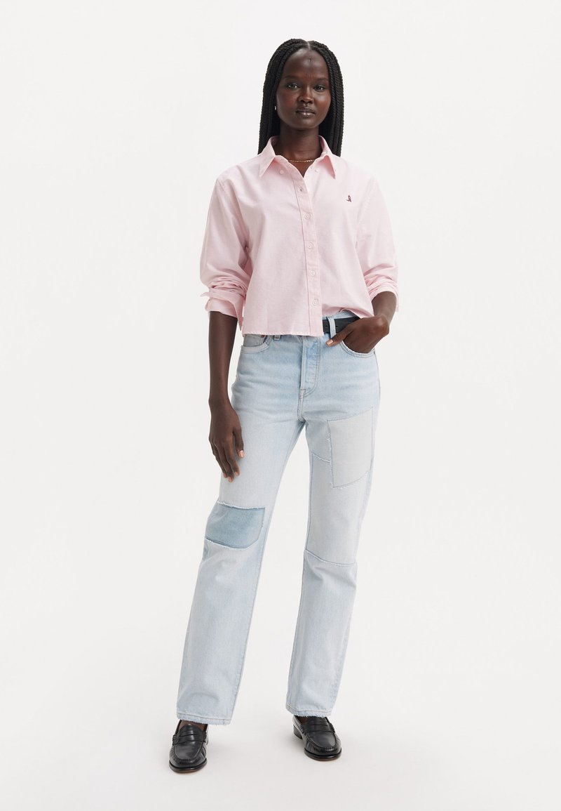 Camicia button-up cropped di un rosa chiaro con maniche lunghe, abbinata a jeans patchwork blu, caratterizzati da pannelli multitexturizzati e una vestibilità rilassata.