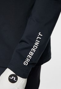 Maglietta a maniche lunghe blu navy con una texture liscia, con scritte bianche lungo la manica. Include un guanto bianco e nero con un logo.