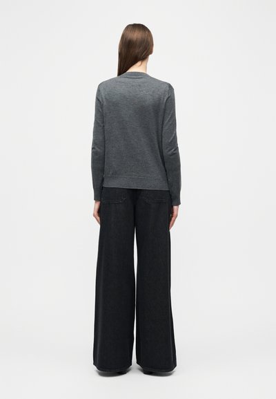 Γυναίκα που κοιτάζει προς τα πίσω, φορώντας ένα σκούρο γκρι πουλόβερ και μαύρα τζιν με φαρδιά μπατζάκια, σε απλό λευκό φόντο.