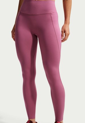 Femme portant des leggings moulants taille haute de couleur mauve, debout, les mains détendues le long du corps sur un fond uni.