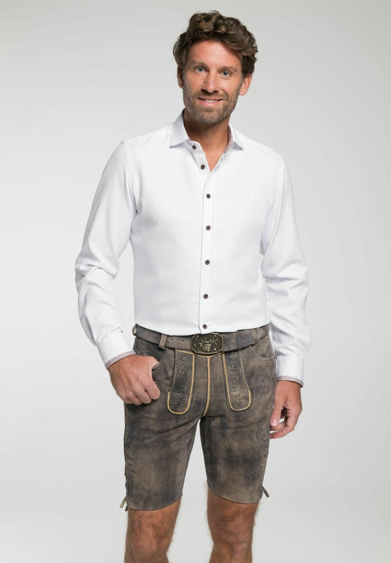 Witte blouse met lange mouwen en bruine knopen, gecombineerd met bruine lederhosen met gouden accenten en bloemige borduursels. Riem met decoratieve gesp.