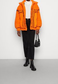 Jasně oranžová nadýchaná bunda se dvěma předními kapsami, černé pruhované detaily, bílý rolák, černé džíny a černé kotníkové boty.