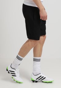 Shorts de sport noirs à coupe décontractée, associés à des chaussures de football blanches avec des rayures noires et des accents verts, ainsi qu'à des chaussettes blanches à rayures.