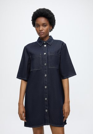 Femme portant une robe chemise en jean bleu foncé à manches courtes avec deux poches poitrine, debout devant un fond clair uni.