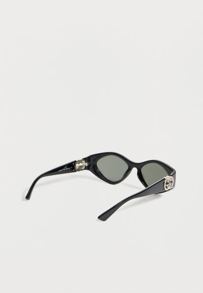 Μαύρα γυαλιά ηλίου τύπου cat-eye με σκοτεινούς φακούς και ασημένια διακοσμητικά λογότυπα στα μπράτσα, τοποθετημένα πάνω σε λευκή επιφάνεια.