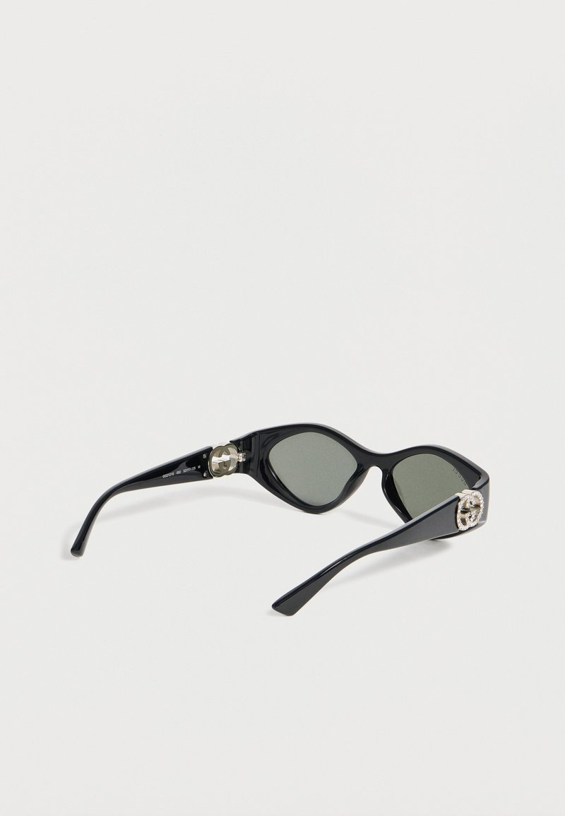 Schwarze Cat-Eye-Sonnenbrille mit dunklen Gläsern und silbernen dekorativen Logos an den Bügeln, auf einer weißen Oberfläche liegend.