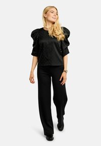 Čierna košeľa s nafúknutými rukávmi a vzorom strieborných bodiek, spárovaná s čiernymi širokými nohavicami a čiernou obuvou. Hladká textúra, jednoduchý dizajn.