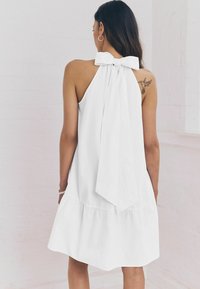 Abito bianco senza maniche con un fiocco sul retro, caratterizzato da un orlo svasato e una texture di tessuto liscia. Design semplice e minimalista.