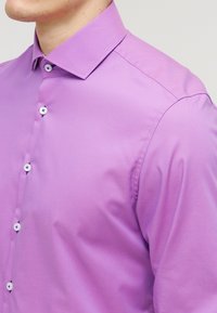 Mężczyzna ubrany w jasnofioletową koszulę na guziki z kołnierzykiem, przyciętą na wysokości szyi i środkowego tułowia, na neutralnym tle.