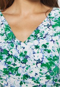 Blommig wrap-top i nyanser av grönt och blått mot en vit bakgrund. Materialet verkar mjukt med en slät textur och har en V-ringning.