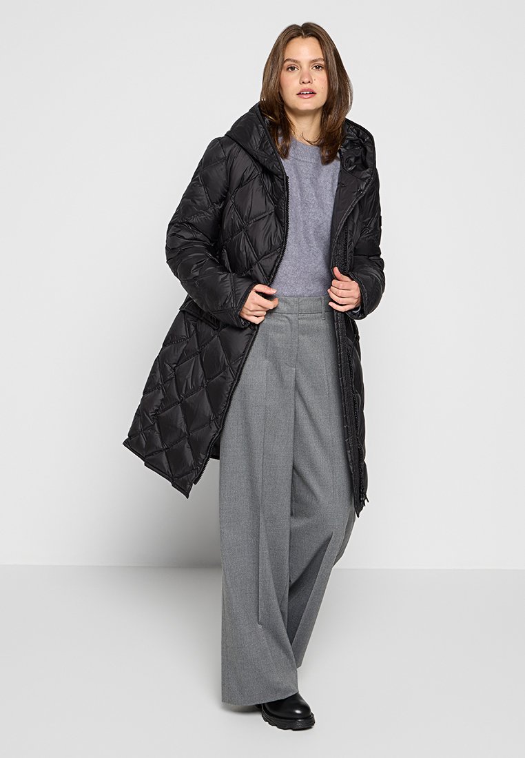 Donna che indossa un lungo cappotto trapuntato nero su un maglione grigio e ampi pantaloni grigi, in piedi di fronte a uno sfondo chiaro e semplice.