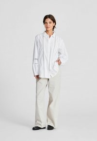 Camicia bianca a maniche lunghe con bordo in pizzo e colletto a camicia. Abbinata a pantaloni beige a gamba larga e scarpe nere. Design semplice nel complesso.