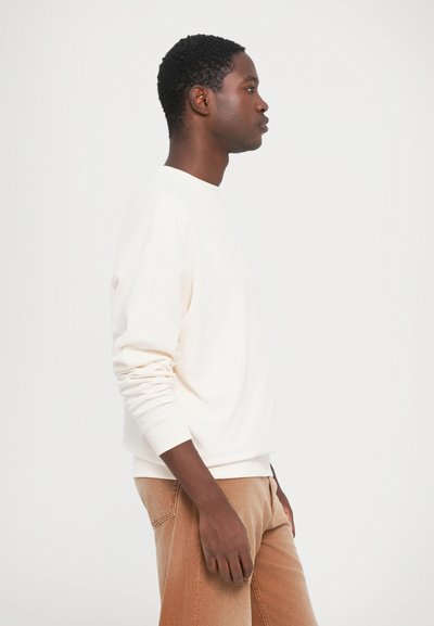 Mann med kort krøllete hår iført en hvit langermet skjorte og lysebrune jeans som står i profil mot en enkel hvit bakgrunn.