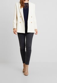 White double-breasted blazer with gold buttons, paired with a navy top and dark gray skinny jeans. Model wears beige ankle boots.