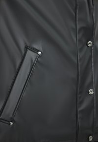 Chaqueta negra impermeable con textura suave, botones de presión y un bolsillo lateral con un acento decorativo de botón plateado.