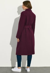 Burgundy takki, jossa on tyylikäs muotoilu, vyöllinen vyötärö, pitkät hihat ja lovikaulus, yhdistettynä tummansinihin legginsseihin ja valkoisiin tennareihin.