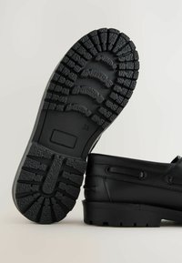 Chaussures en cuir noir avec une semelle en caoutchouc texturé, dotée de profondeurs de relief et d'un talon plat. Coutures visibles et œillets pour les lacets.