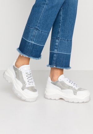 Person iført blå jeans med frynset kant og hvide chunky sneakers med grå meshdetaljer, stående på en lysegrå gulv.