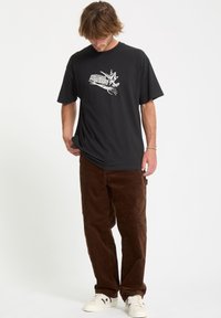 Czarny t-shirt bawełniany z białym nadrukiem z przodu, w połączeniu z brązowymi spodniami sztruksowymi i białymi trampkami.