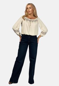 Kremowa bluzka z koronkowym wykończeniem wzdłuż dekoltu i rękawów, połączona z ciemnoniebieskimi dżinsami o wysokim stanie i szerokiej nogawce.