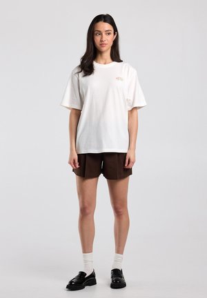 Jeune femme aux longs cheveux foncés portant un t-shirt blanc oversize, un short marron, des chaussettes blanches et des mocassins noirs, debout devant un fond uni.