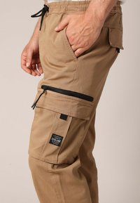 Pantalon cargo kaki en coton résistant, avec une poche zippée, une poche latérale et une ceinture à cordon de serrage réglable. Gros plan sur la jambe.